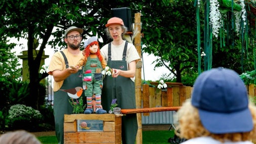 A photography of two puppeteers manipulating a small puppet on a table in an outside setting with audience watching. They are all wearing green dungarees and a peak cap.